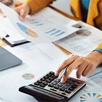 Person using a calculator while reviewing financial reports and charts at a desk.