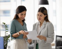 Two women reviewing joint project details, discussing commercial proposal standing in modern office, verifying printed documents or contracts, collaborating on creative ideas, planning upcoming tasks