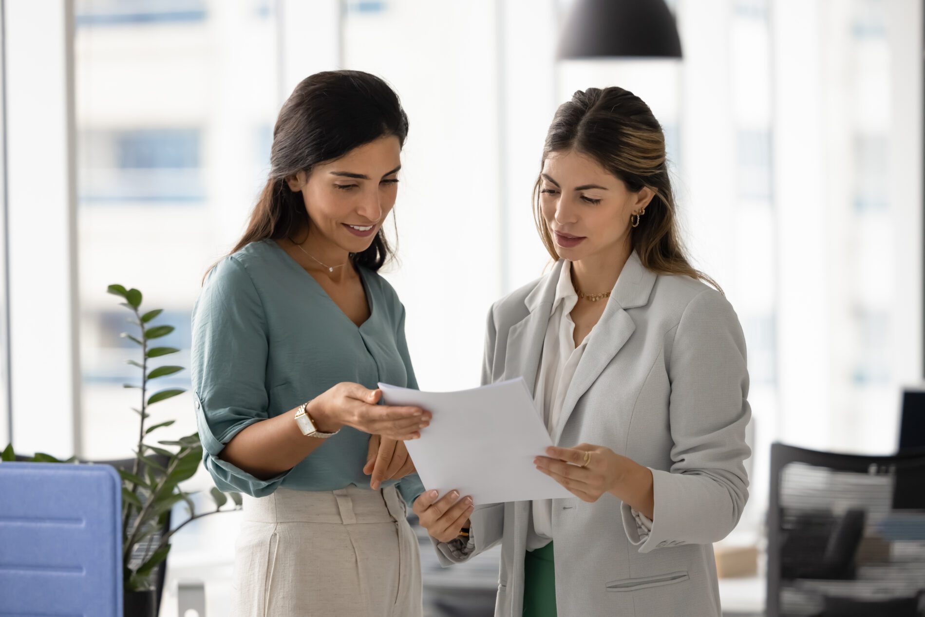 Two women reviewing joint project details, discussing commercial proposal standing in modern office, verifying printed documents or contracts, collaborating on creative ideas, planning upcoming tasks