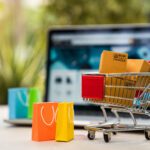 Product package boxes in cart with shopping bag and laptop computer with blurred web store shop on screen for online shopping and delivery concept